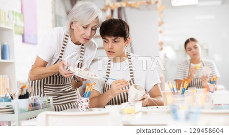 Elderly woman assisting adult son painting ceramic mugs in family workshop 129458604