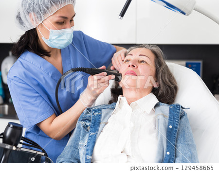Female doctor cosmetologist performing machine facial procedure to mature woman in clinic Female doctor cosmetologist performing machine facial procedure to mature woman in clinic 129458665