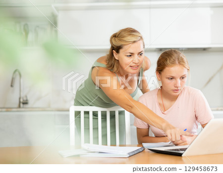 Mother looks at laptop and prompts and helps teen girl to write in notebook to complete task Mother looks at laptop and prompts and helps teen girl to write in notebook to complete task 129458673