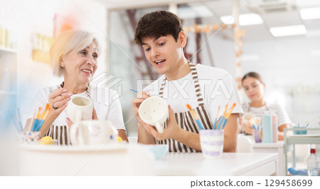 Cheerful elderly woman and guy painting ceramic mugs during pottery masterclass Cheerful elderly woman and guy painting ceramic mugs during pottery masterclass 129458699