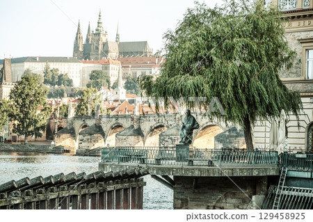 The statue of composer Bedrich Smetana stands by the Vltava River, offering a stunning view of Prague's architecture, including the impressive castle and iconic Charles Bridge, bathed in sunlight. 129458925