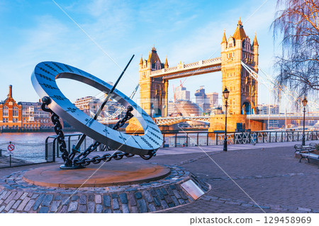A large sundial stands prominently by the Thames River, with Tower Bridge in the background. The sun casts shadows on the sundial, marking the time while tourists enjoy the view. 129458969