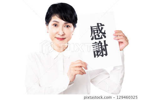 一位身穿白襯衫的老年婦女微笑著,手裡拿著一張寫著「謝謝」的紙條 一位身穿白襯衫的老年婦女微笑著,手裡拿著一張寫著「謝謝」的紙條 129460325