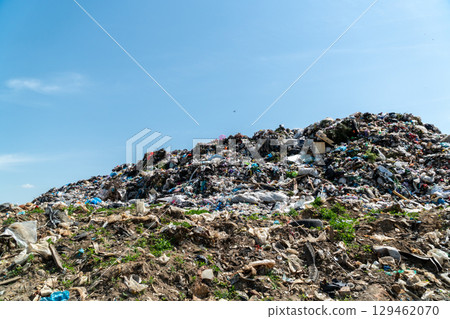 A massive pile of trash dominates the landscape, surrounded by scattered debris and greenery under a bright sky A massive pile of trash dominates the landscape, surrounded by scattered debris and greenery under a bright sky 129462070