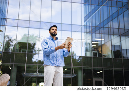 A man in a blue shirt stands in front of a modern glass building, holding a tablet, looking up. A man in a blue shirt stands in front of a modern glass building, holding a tablet, looking up. 129462985
