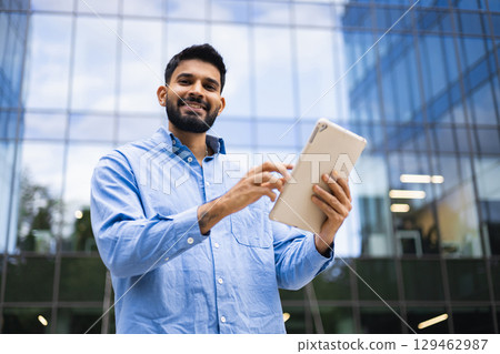 A smiling businessman of Indian descent uses a tablet in front of a modern glass building, conveying a sense of professional success and technological integration. A smiling businessman of Indian descent uses a tablet in front of a modern glass building, conveying a sense of professional success and technological integration. 129462987