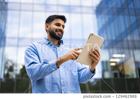 A smiling man in a blue shirt uses a tablet outside of a modern building. A smiling man in a blue shirt uses a tablet outside of a modern building. 129462988