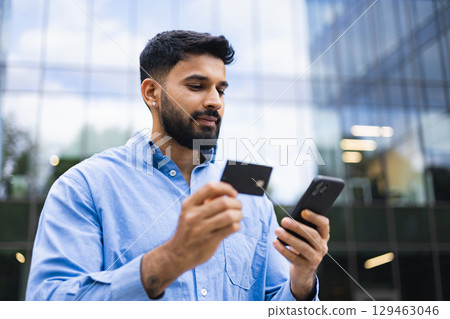 An Indian man in a blue shirt is shown holding a credit card and a smartphone in front of a modern building. An Indian man in a blue shirt is shown holding a credit card and a smartphone in front of a modern building. 129463046