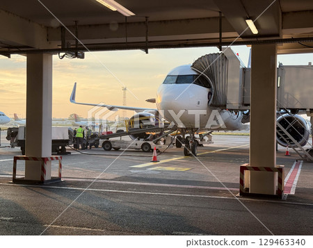 Passenger airplane connected to boarding bridge at airport during golden hour. Travel infrastructure, global mobility, and departure preparation at sunrise. 129463340