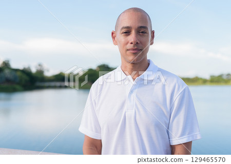 Confident bald man in white polo shirt posing by lake in serene outdoor portrait Confident bald man in white polo shirt posing by lake in serene outdoor portrait 129465570
