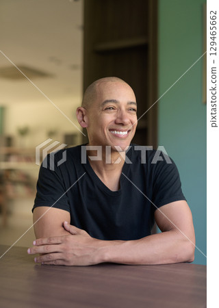 Smiling hispanic man in library wearing black t-shirt looking through window indoors 129465662