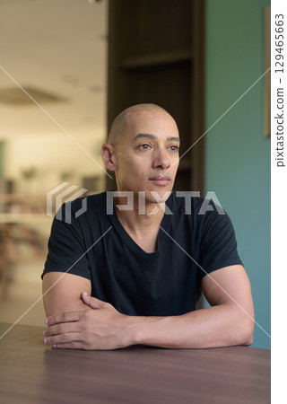 Serious Hispanic bald man in library wearing black t-shirt looking through window indoors 129465663