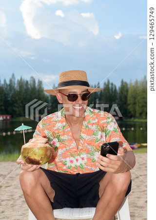 Man tourist sitting on beach using mobile phone and holding coconut water Man tourist sitting on beach using mobile phone and holding coconut water 129465749
