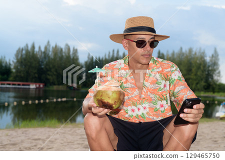 Man tourist sitting on beach using mobile phone and holding coconut water Man tourist sitting on beach using mobile phone and holding coconut water 129465750
