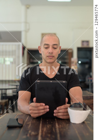 Bald Hispanic man sitting in outdoors cafe restaurant using digital tablet computer Bald Hispanic man sitting in outdoors cafe restaurant using digital tablet computer 129465774