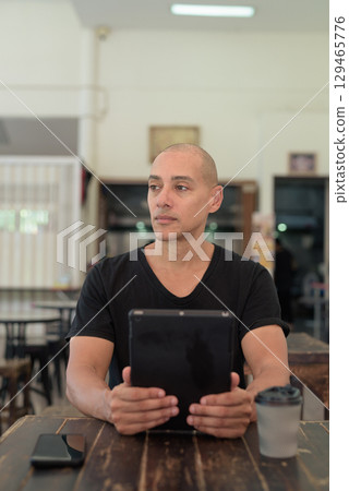 Bald Hispanic man sitting in outdoors cafe restaurant using digital tablet computer Bald Hispanic man sitting in outdoors cafe restaurant using digital tablet computer 129465776