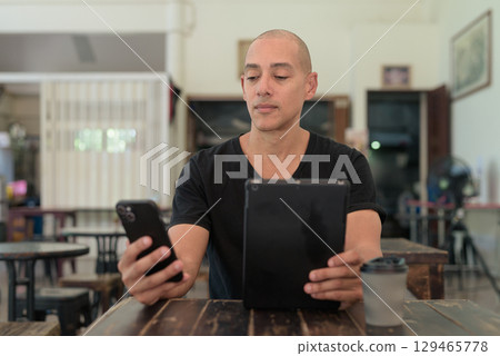 Bald Hispanic man sitting in outdoors cafe restaurant using phone and digital tablet computer 129465778