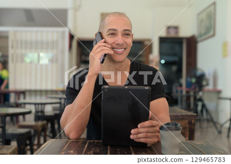 Bald Hispanic man sitting in outdoors cafe restaurant using phone and digital tablet computer 129465783