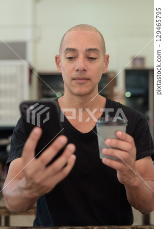 Bald man in black t-shirt sitting in outdoors cafe restaurant using phone and holding coffee cup 129465795