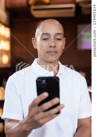 Confident Bald Hispanic man in white polo shirt sitting in Chinese style coffee shop using phone 129465814