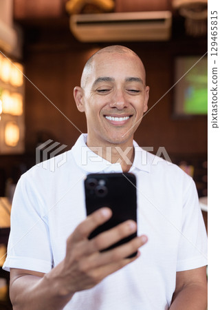 Confident Bald Hispanic man in white polo shirt sitting in Chinese style coffee shop using phone 129465815