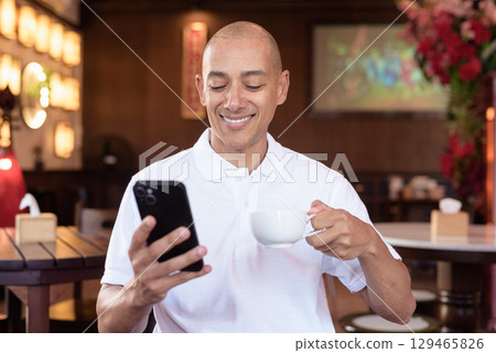 Confident Bald Hispanic man in white polo shirt sitting in Chinese style coffee shop using phone 129465826