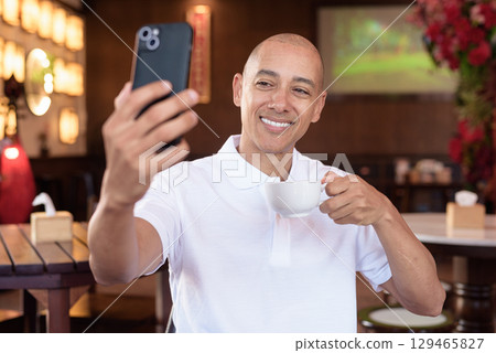 Confident Bald Hispanic man in white polo shirt sitting in Chinese style coffee shop using phone 129465827
