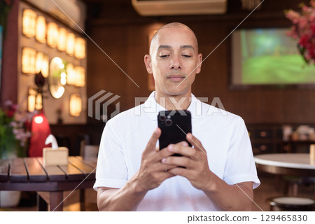 Confident Bald Hispanic man in white polo shirt sitting in Chinese style coffee shop using phone 129465830