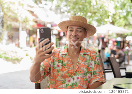 Hispanic man using phone taking selfie at outdoor cafe wearing Panama hat and short sleeve shirt 129465871