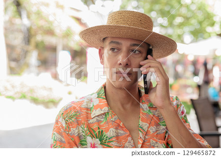 Hispanic man talking on phone at outdoor cafe wearing Panama hat and short sleeve shirt 129465872