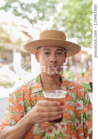 Hispanic man holding iced coffee at outdoor cafe wearing Panama hat and short sleeve shirt 129465880