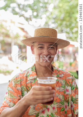 Hispanic man holding iced coffee at outdoor cafe wearing Panama hat and short sleeve shirt 129465881