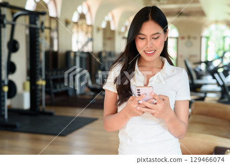Young Asian woman in polo shirt at gym using phone in modern indoor training location 129466074