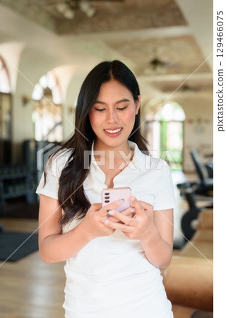 Young Asian woman in polo shirt at gym using phone in modern indoor training location 129466075