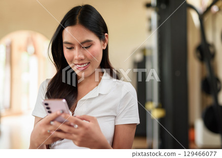 Young Asian woman in polo shirt at gym using phone in modern indoor training location 129466077