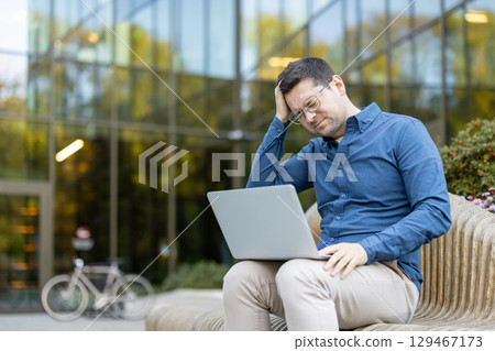 A concerned young man holding his head while working on a laptop outdoors in a modern setting. The image conveys stress, focus, and challenges associated with business work or problem-solving. 129467173