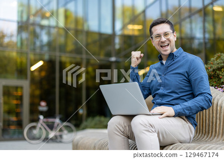Smiling man sitting outside with a laptop celebrating success, conveying joy and positivity. Smiling man sitting outside with a laptop celebrating success, conveying joy and positivity. 129467174