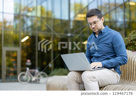 Professional individual focused on work using a laptop outside a contemporary glass building, depicting productivity in a serene outdoor setting. Professional individual focused on work using a laptop outside a contemporary glass building, depicting productivity in a serene outdoor setting. 129467176
