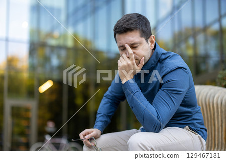 A man is seated outside near a modern building, appearing thoughtful and stressed. He holds his glasses and seems to ponder or feel concerned about a situation. Reflections present on the glass. A man is seated outside near a modern building, appearing thoughtful and stressed. He holds his glasses and seems to ponder or feel concerned about a situation. Reflections present on the glass. 129467181