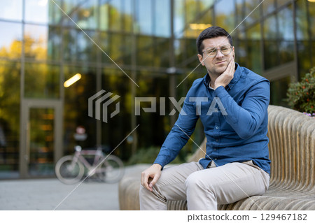 A man in casual attire sitting outdoors in front of a glass building, expressing discomfort by holding his face, likely due to dental pain. 129467182