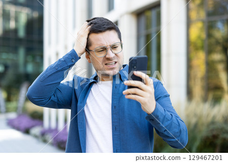 A man looks frustrated while using his smartphone outdoors. The building backdrop and his expression suggest a displeasing or confusing interaction with technology or communication. 129467201