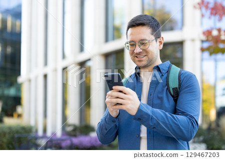 A cheerful individual interacts with a smartphone while standing in front of a contemporary building. The image conveys positivity, modern communication 129467203