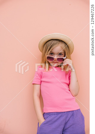 Young girl in stylish outfit and sunglasses poses confidently against pink wall background Young girl in stylish outfit and sunglasses poses confidently against pink wall background 129467226