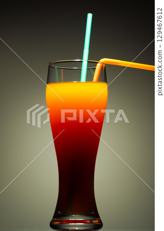 A tall glass containing a layered beverage, likely a Tequila Sunrise cocktail, against a dark, muted background 129467612