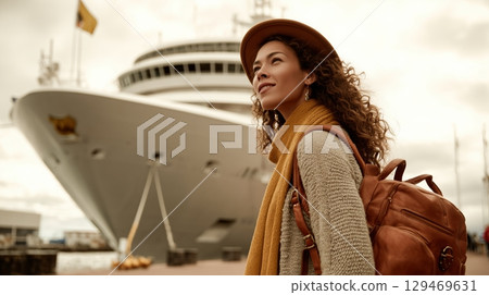 Young woman with curly hair wearing hat and scarf, standing near large cruise ship at harbor 129469631