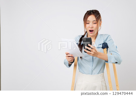 Injured woman with crutches holds mobile phone and bill paper, confused by calculator numbers. Crutches help her stand as she struggles with payment problems. isolated studio white background Injured woman with crutches holds mobile phone and bill paper, confused by calculator numbers. Crutches help her stand as she struggles with payment problems. isolated studio white background 129470444