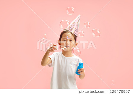 Charming Chinese Female Child in Conical Party Hat Making Soap Bubbles Cheerfully Standing on Pink Isolated Background 129470997