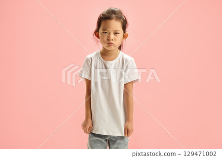 Angry Little Asian Girl Having Gloomy Expression on Face Resentment Looking to Camera Does not Agree with Someone's Opinion, Defiantly Standing in Stubborn Pose Expressed in Body Language against pink 129471002
