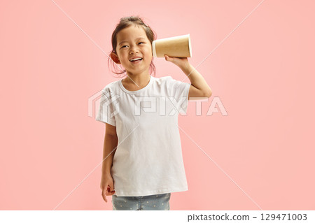 Cheerful cute Asian child girl spying using paper cup for listening, trying overheal someone, holds hand with cup near ear to enhance the sound, wearing in neutral clean white t shirt on pink isolated 129471003