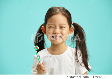 Cute Asian ethnicity child girl with a two ponytails holding toothbrush and happy smiling preparing brush self teeth standing over blue isolated background. Children health care concept. Cute Asian ethnicity child girl with a two ponytails holding toothbrush and happy smiling preparing brush self teeth standing over blue isolated background. Children health care concept. 129471008